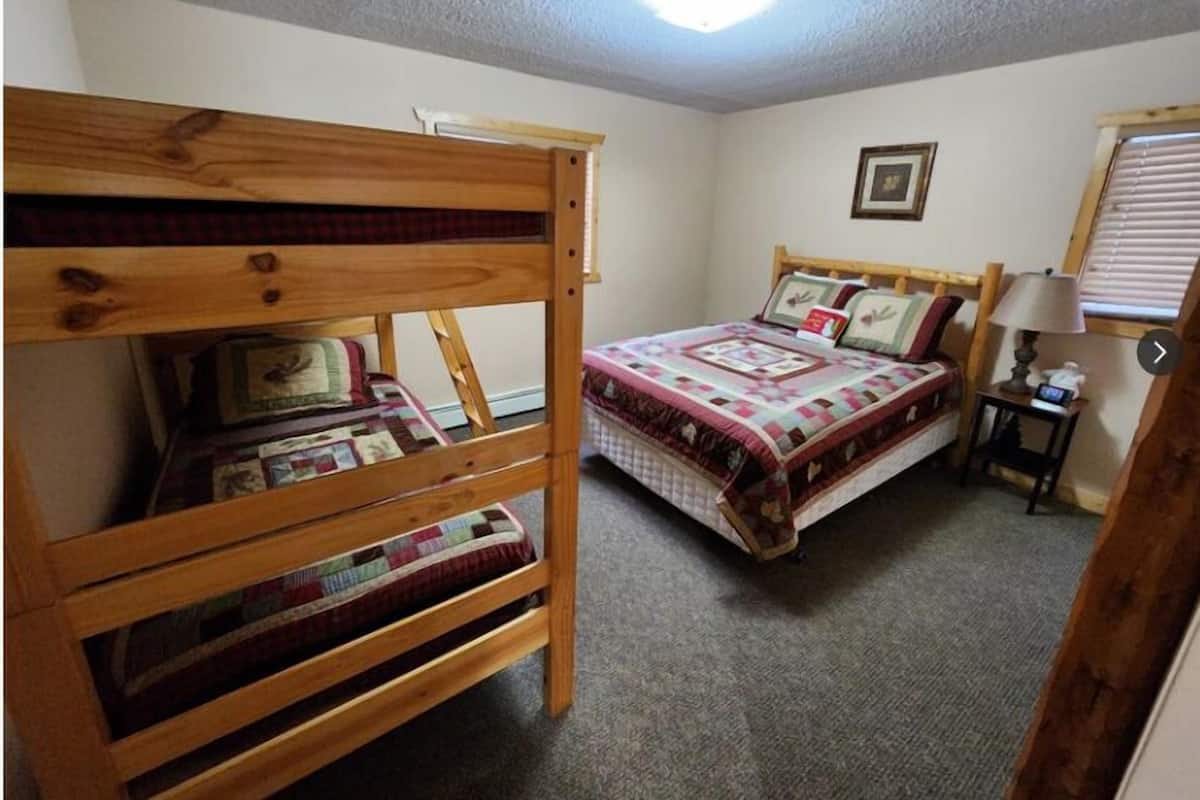 Un dormitorio con una cama litera y una cama individual, ambas con colchas. Hay un cuadro en la pared y una lámpara sobre una mesita de noche.