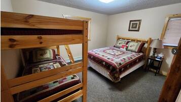 A bedroom with a bunk bed and a single bed, both with quilts. There is a framed picture on the wall and a lamp on a bedside table.