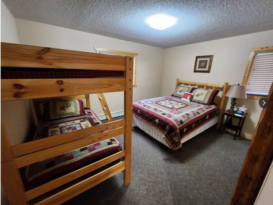A bedroom with a bunk bed and a single bed, both with quilts. There is a framed picture on the wall and a lamp on a bedside table.