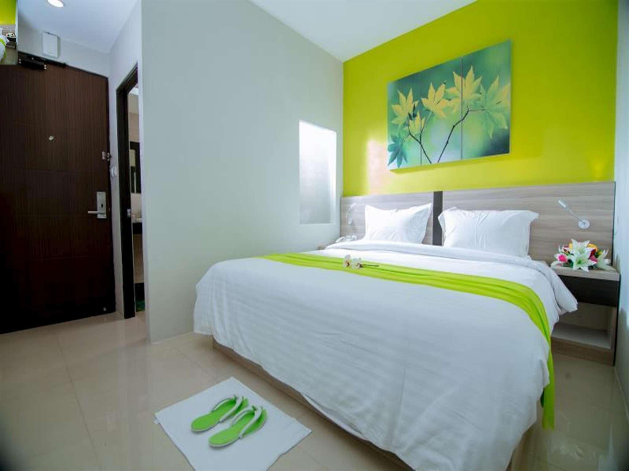 A hotel room with a bed, a nightstand, a painting on the wall, and a pair of slippers on the floor.