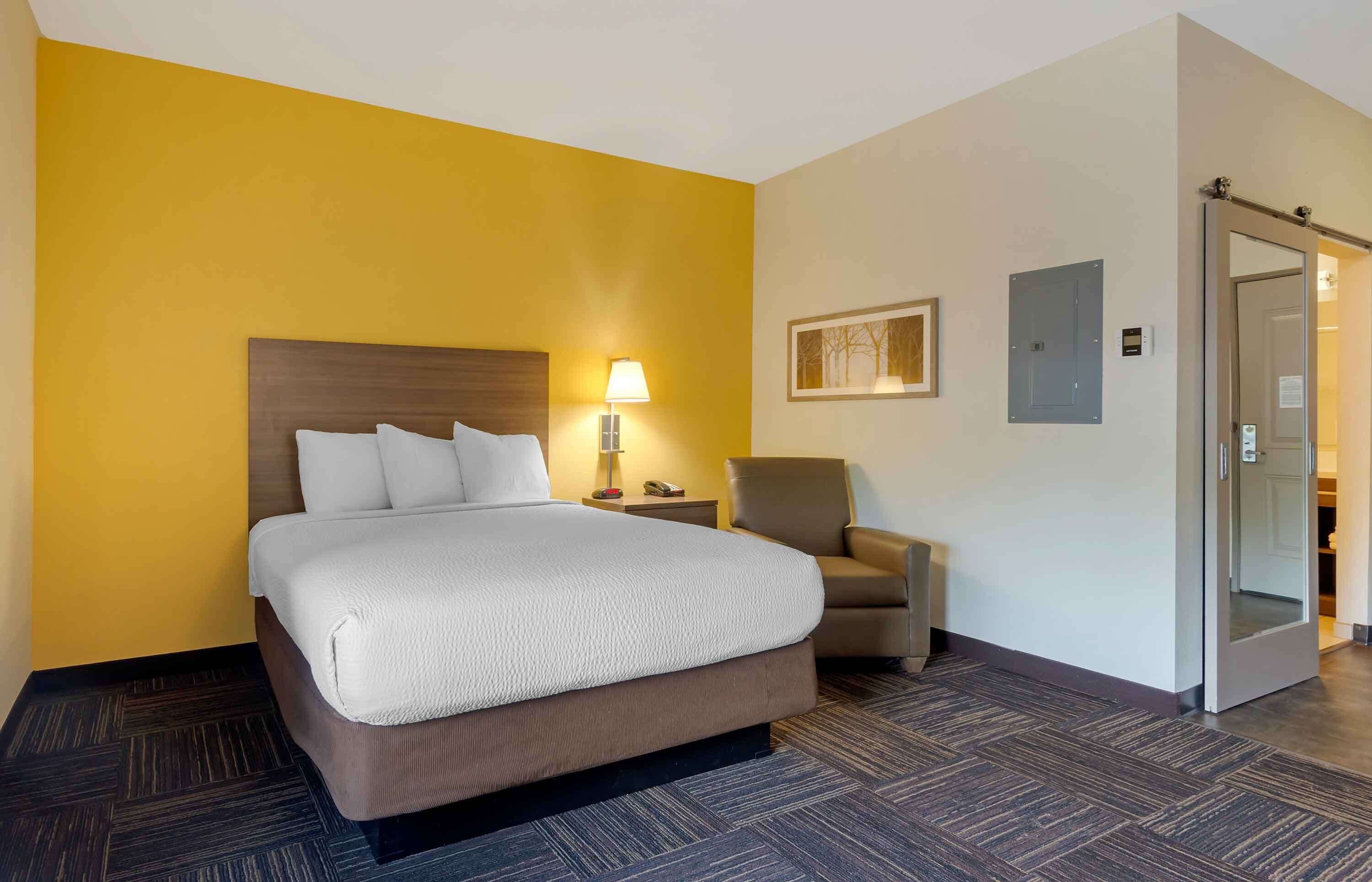 Una habitación de hotel con una cama grande, una lámpara de noche, una silla y una puerta que da a otra habitación.