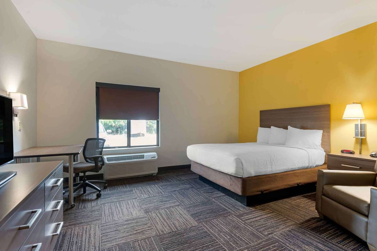 Een hotelkamer met een bed, bureau, stoel, televisie en een raam met jaloezieën.