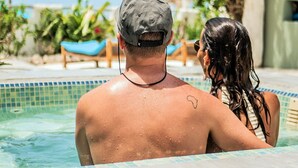 A couple in a swimming pool with a tropical setting, including palm trees and a thatched-roof structure.