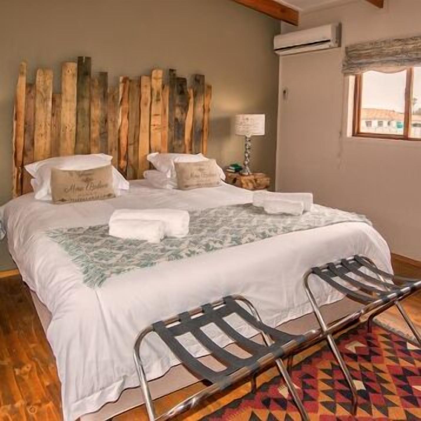 Una cama con sábanas blancas, dos almohadas y una manta. Tiene un cabecero de madera, una mesita de noche con una lámpara y una ventana con vistas.