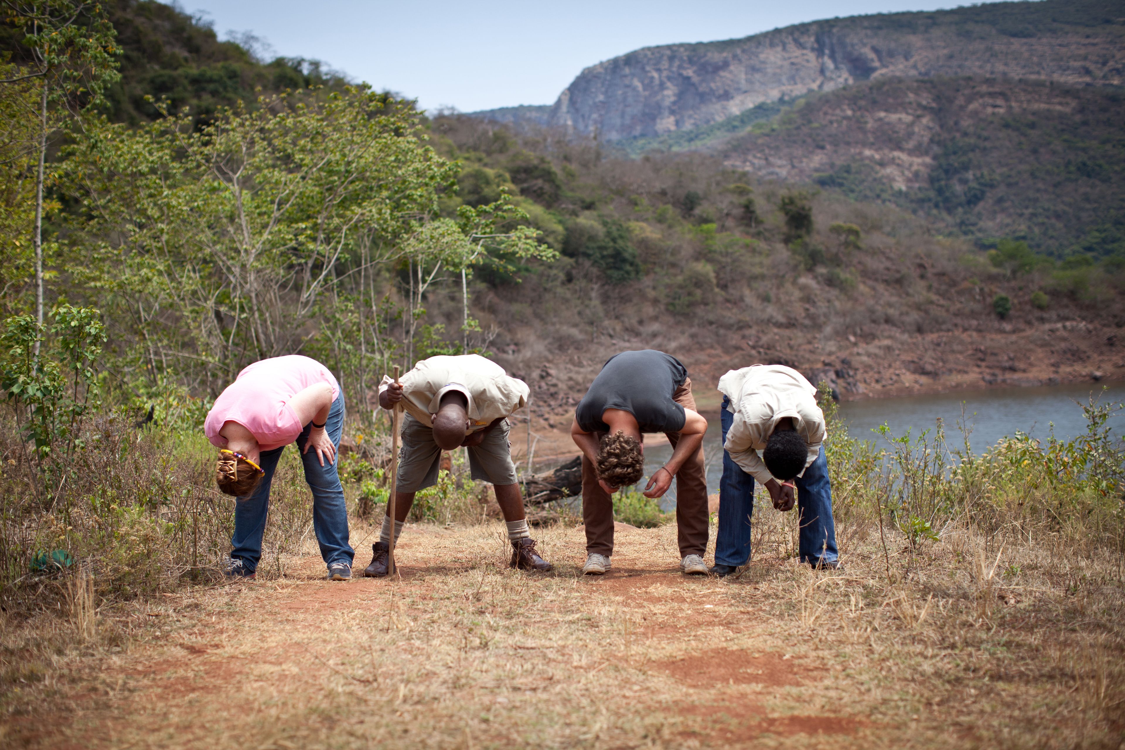 Four people bending over a dirt path in a natural setting with a lake and hills in the background.