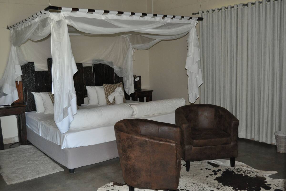 A bedroom with a four-poster bed, a brown armchair, a white sofa, and a large window with curtains.