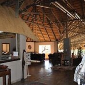 A spacious room with a high ceiling and exposed beams, a large window, and a seating area with a table and chairs. - Shametu River Lodge (Divundu)