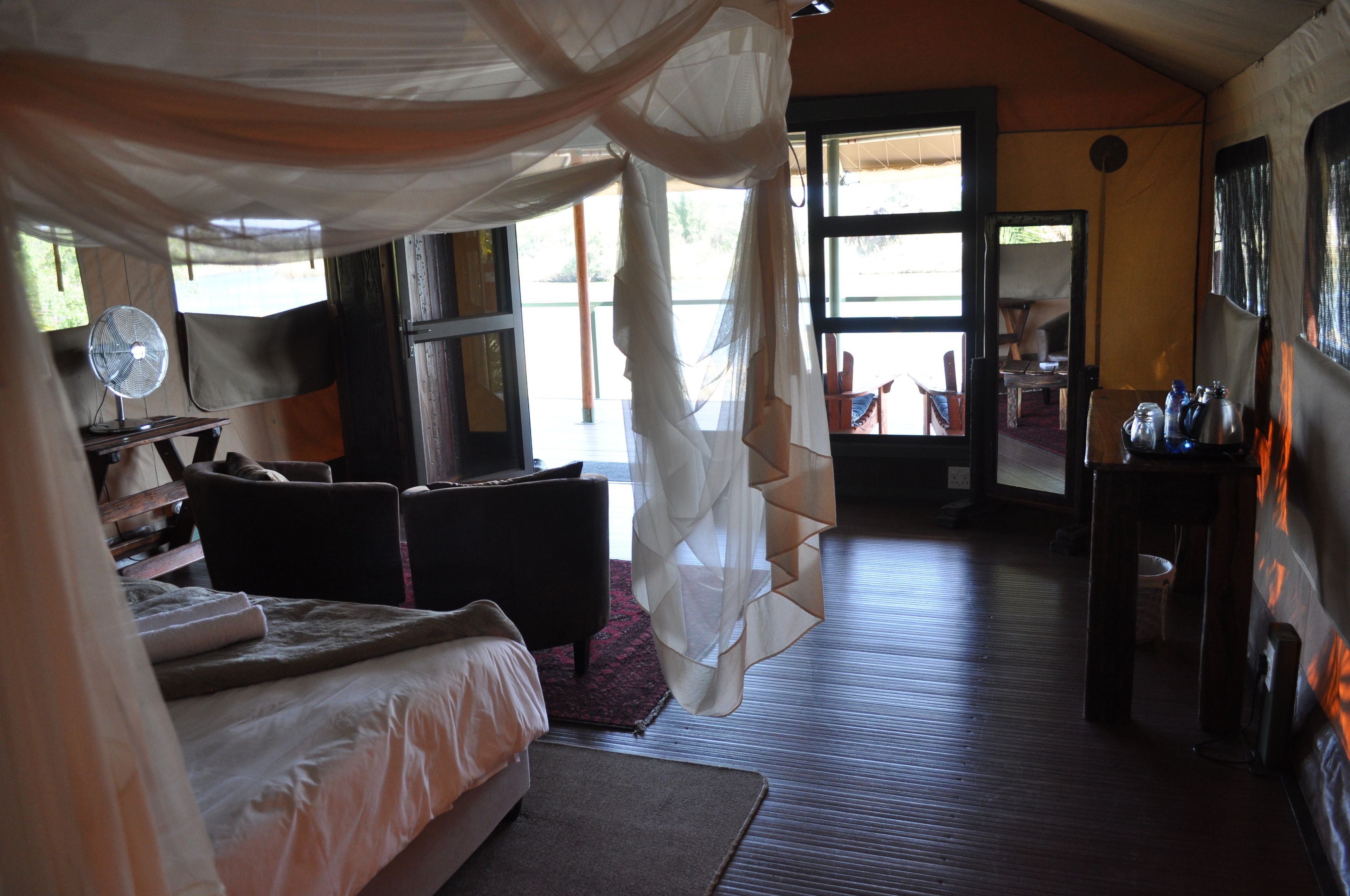A bedroom with a bed, bedside table, and a fan. There is a window with curtains and a view of the outside.
