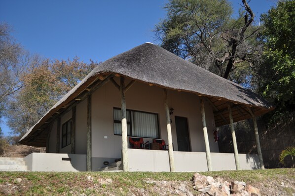 A thatched-roof building with a balcony, surrounded by trees and a rocky landscape.