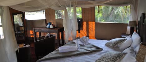 A room with a bed, a desk, and a chair. There is a window with curtains and a view of greenery outside.