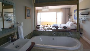 A bathroom with a large bathtub, a sink, a mirror, and a window with a view of a hotel room. - Dolphin Inn Guest House (Cape Town)