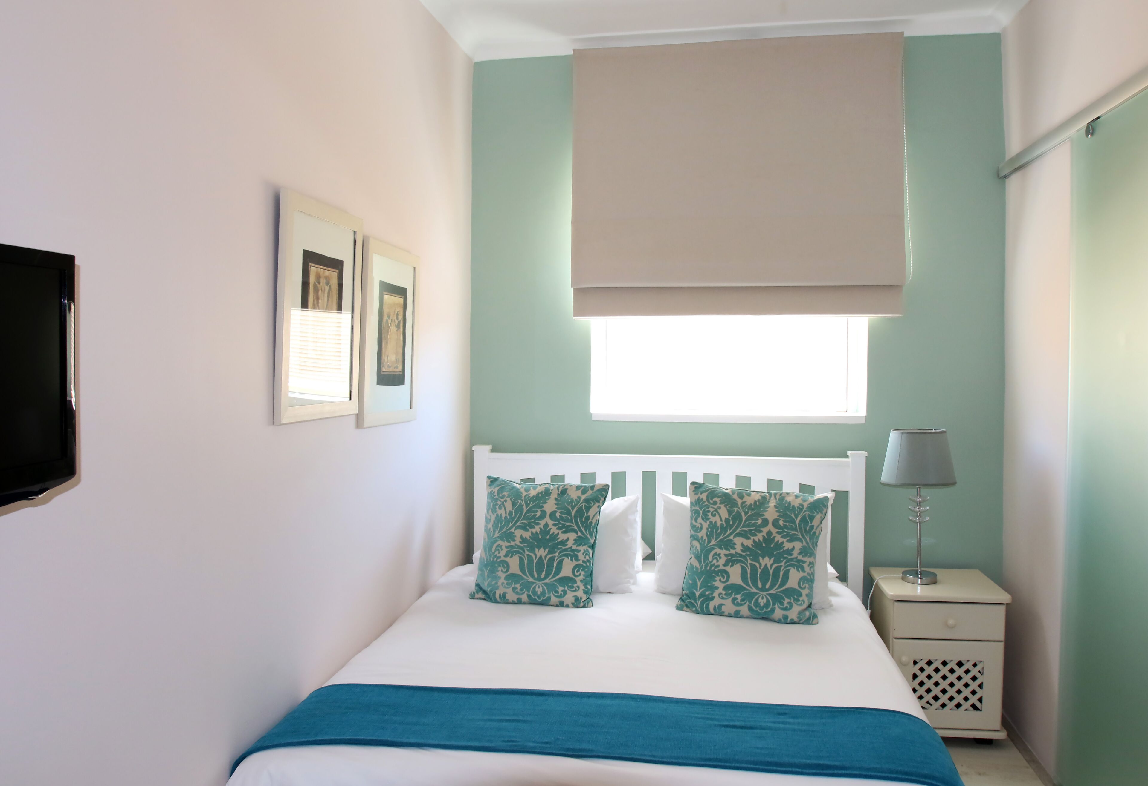 Une chambre bien rangée, avec un lit, une table de chevet sur laquelle se trouve une lampe, une fenêtre avec un store et des tableaux encadrés accrochés au mur.