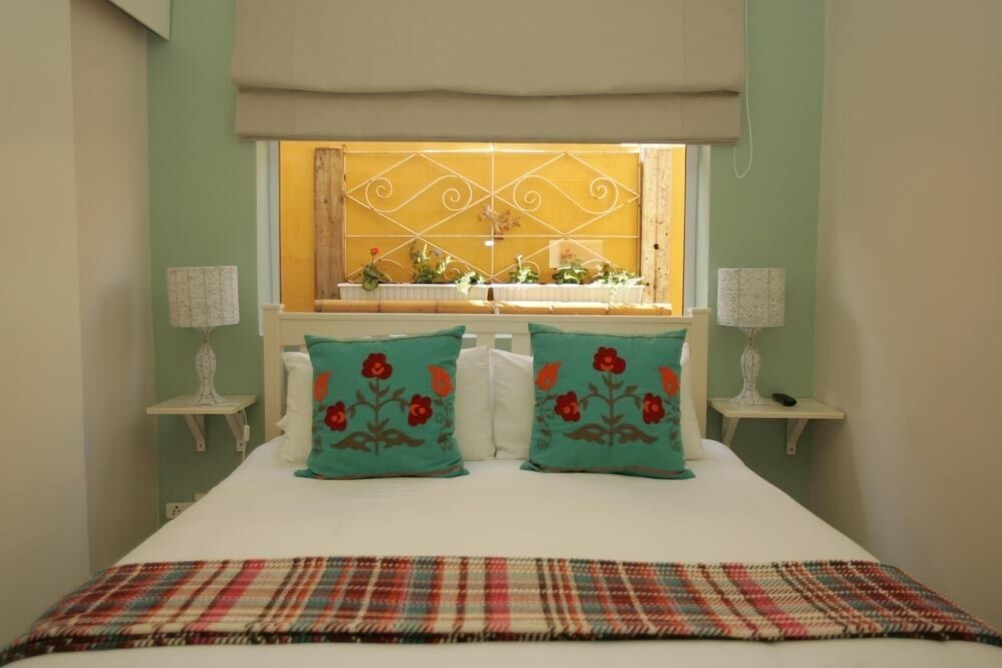 Een slaapkamer met een bed, twee sierkussens met bloemenmotieven, een geruite plaid, nachtkastjes met lampen en een raam met gordijnen.