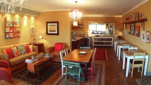 A cozy living room with a wooden dining table, a sofa, and a chandelier. There is a kitchen area in the background with a bar and chairs. - Dolphin Inn Guest House (Cape Town)