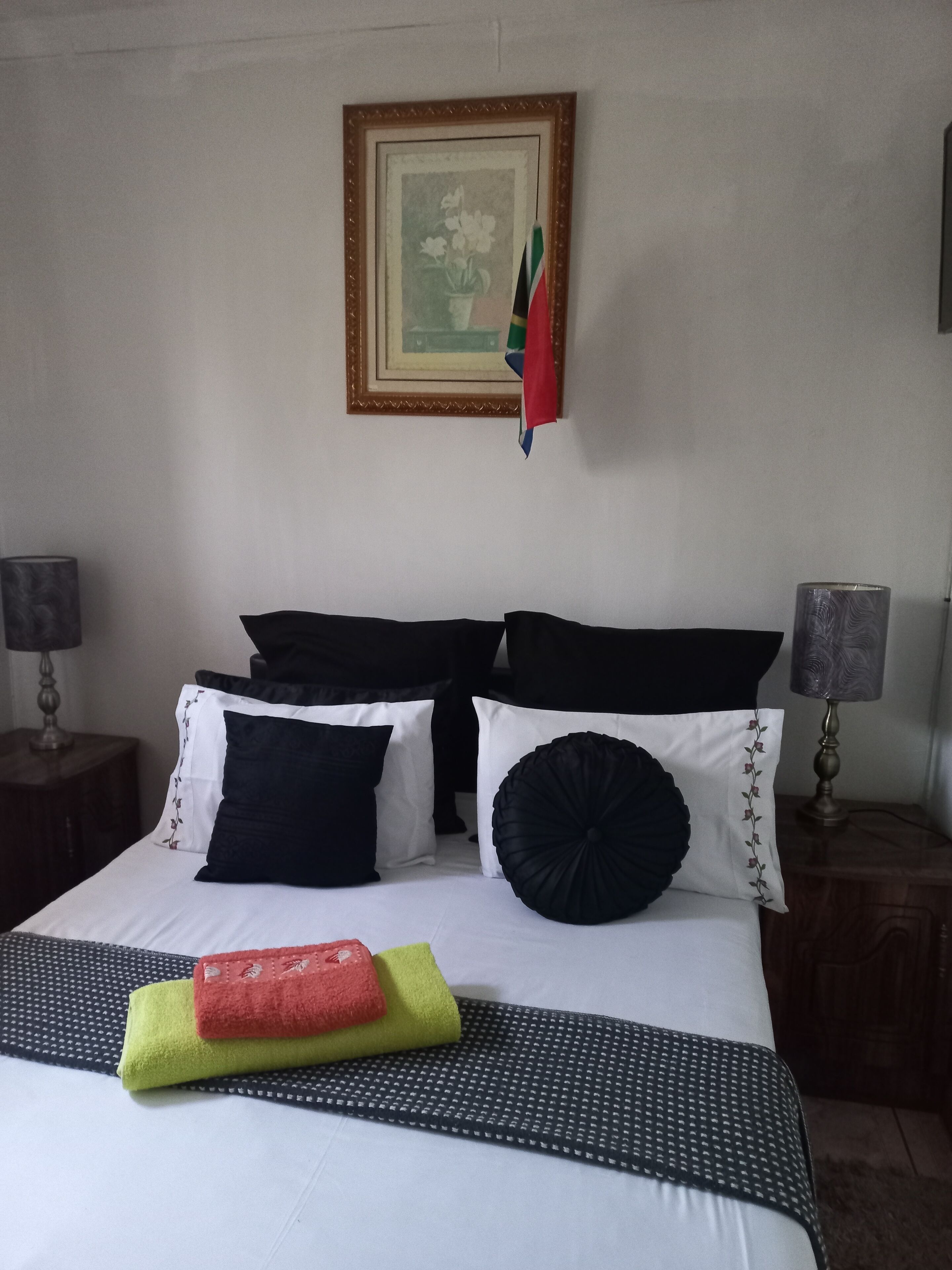 Una cama bien hecha con edredón blanco, almohadas negras y blancas, y una almohada decorativa negra. Hay un cuadro en la pared, una lámpara en la mesita de noche y una toalla sobre la cama.