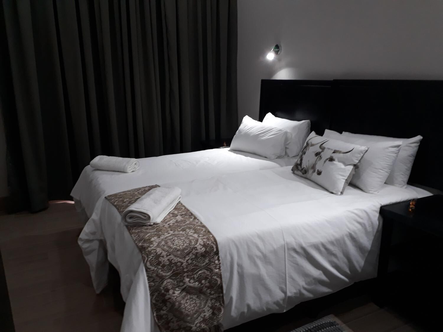 Un lit bien fait, avec du linge de lit blanc et un plaid à motifs, une tête de lit sombre et un luminaire fixé au mur.