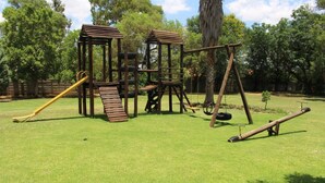 A playground with wooden structures, including a slide and swings, set in a grassy area with trees in the background. - The Willow Tree Guest House (Klerksdorp)