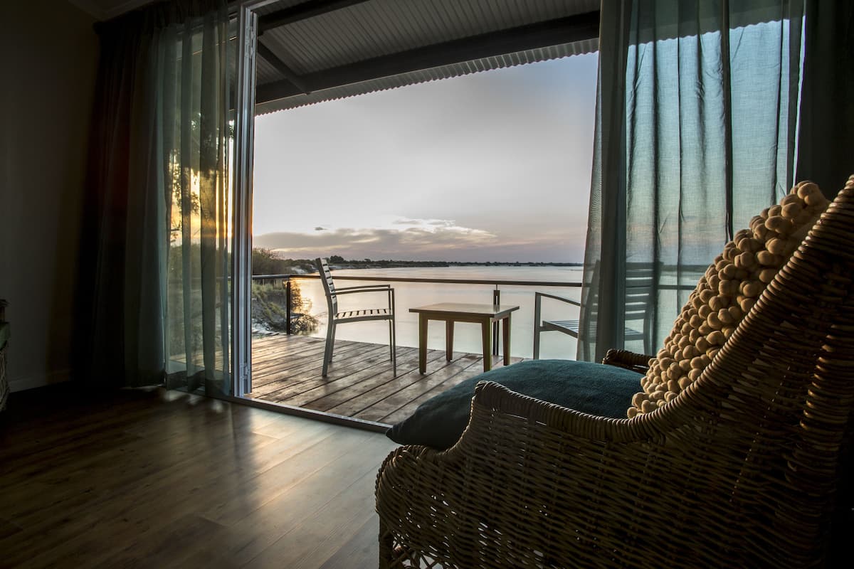 A room with a wicker chair, a small table, and a view of a body of water through large windows.