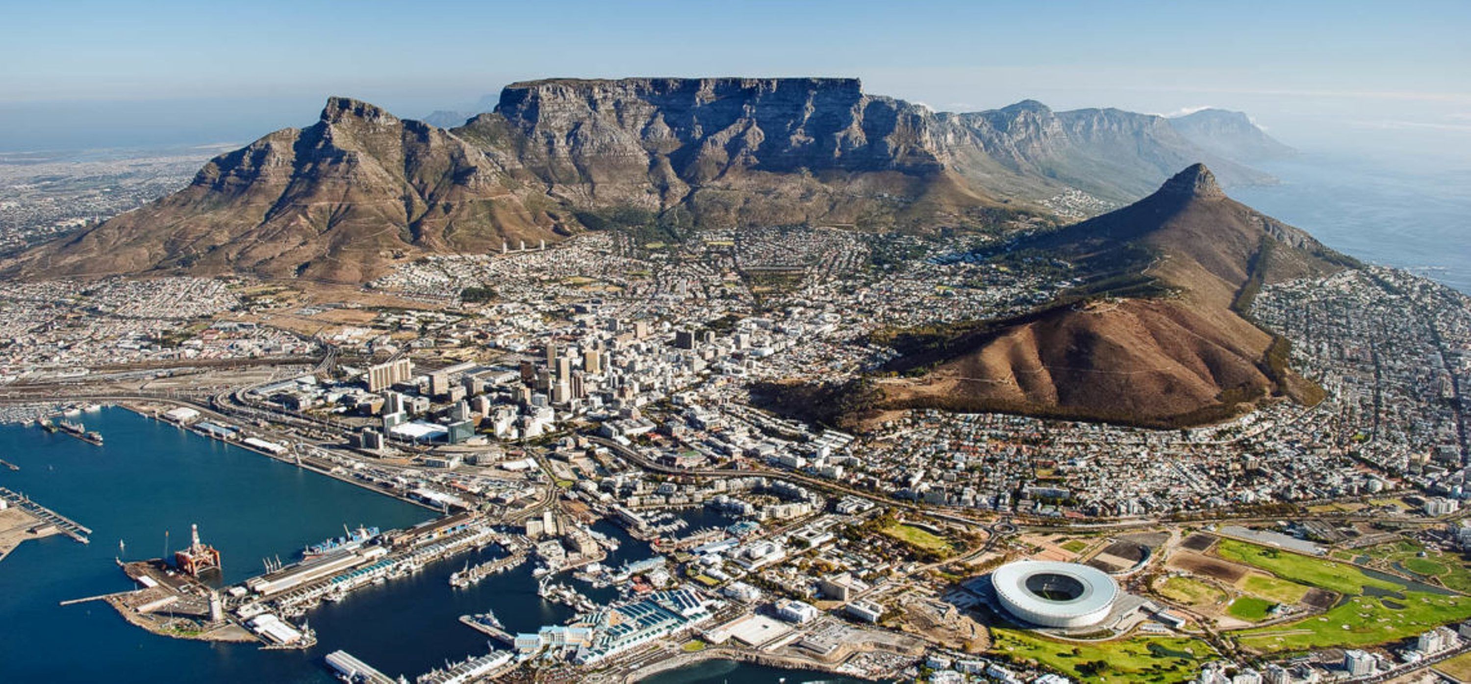 An aerial view of a coastal city with a prominent mountain, a marina, and a stadium.