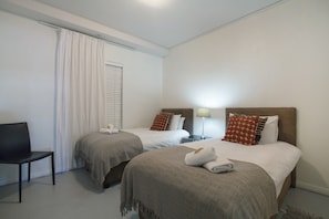 Habitación de hotel con dos camas, una silla, una mesita de noche con lámpara y una ventana con persianas.