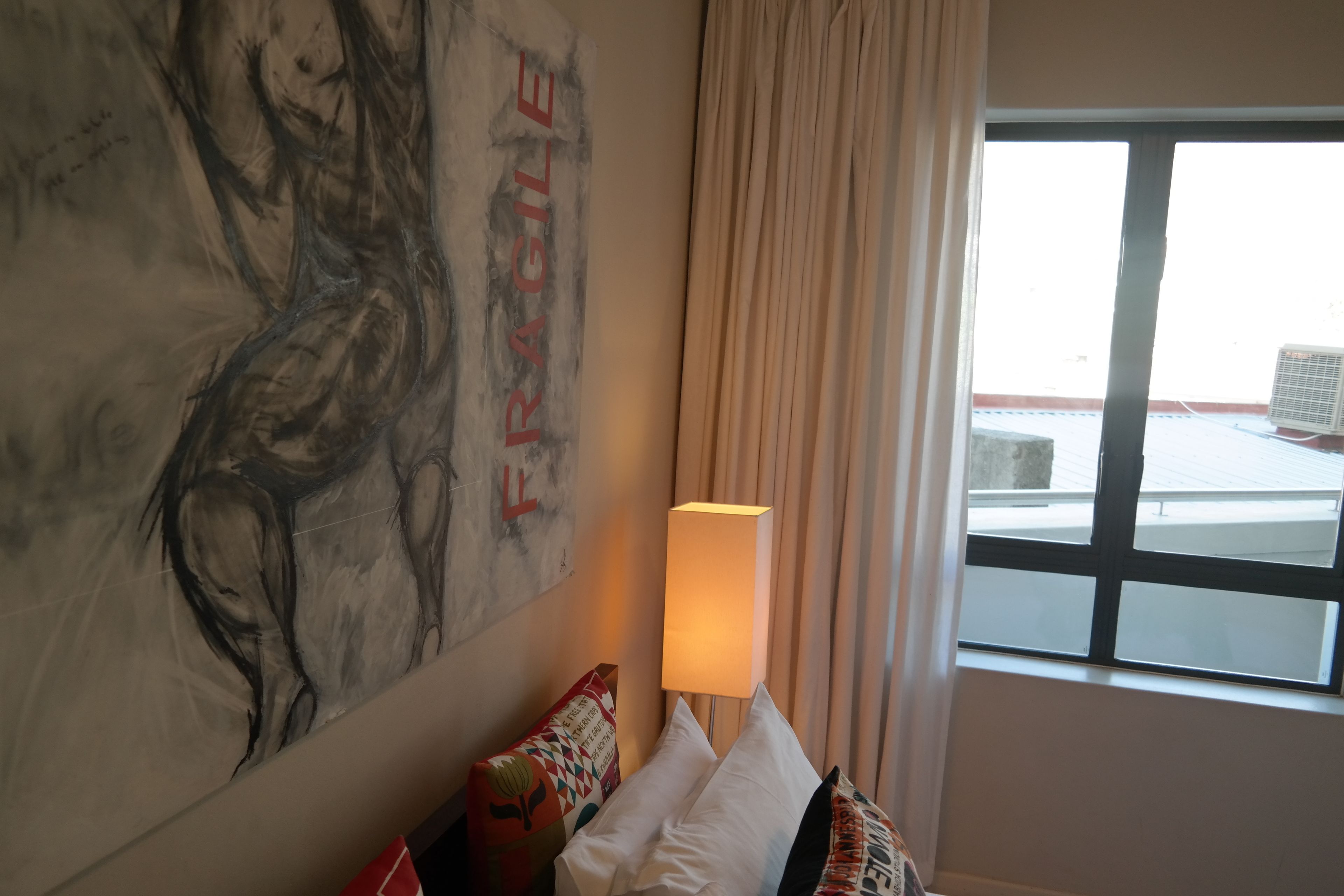 A hotel room with a large abstract painting on the wall, a window with a view of the outside, and a lamp on the bedside table.