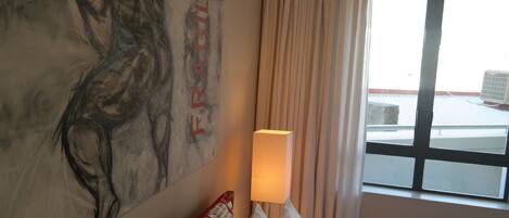 A hotel room with a large abstract painting on the wall, a window with a view of the outside, and a lamp on the bedside table.