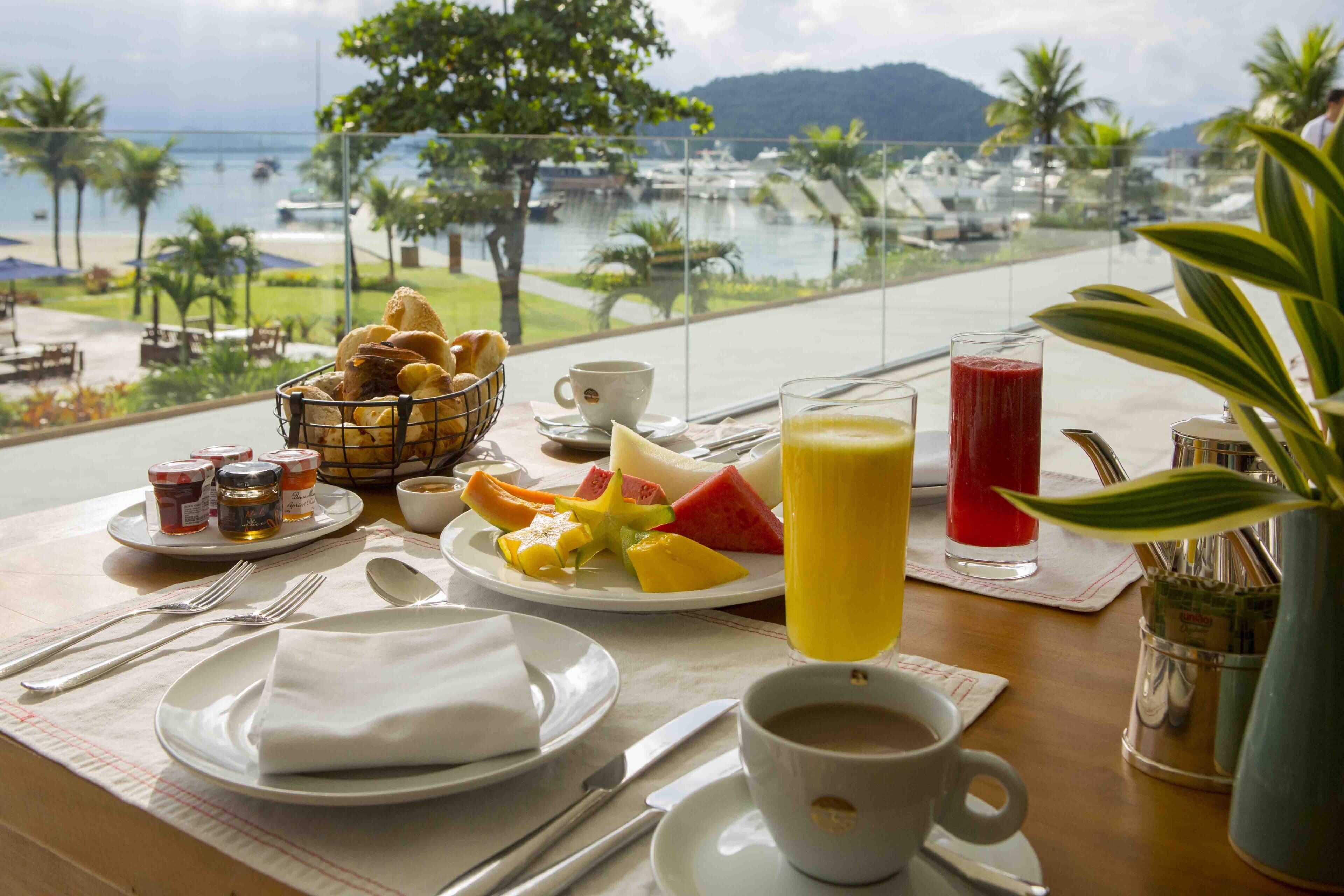 A breakfast table with croissants, fruit, and beverages, overlooking a marina and beach.