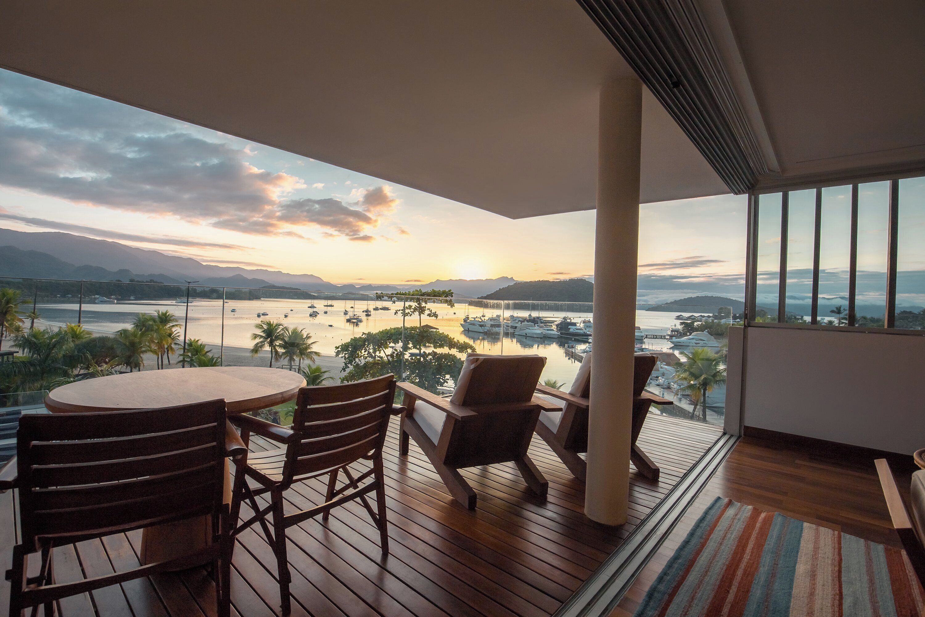 A balcony with a wooden table and chairs, overlooking a marina with boats and a sunset view.