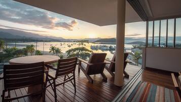 A balcony with a wooden table and chairs, overlooking a marina with boats and a sunset view.