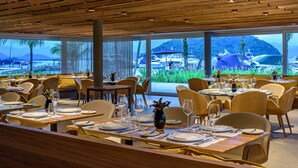 A restaurant with wooden ceiling and large windows offering a view of boats and greenery. Tables are set with white tablecloths and wine glasses.