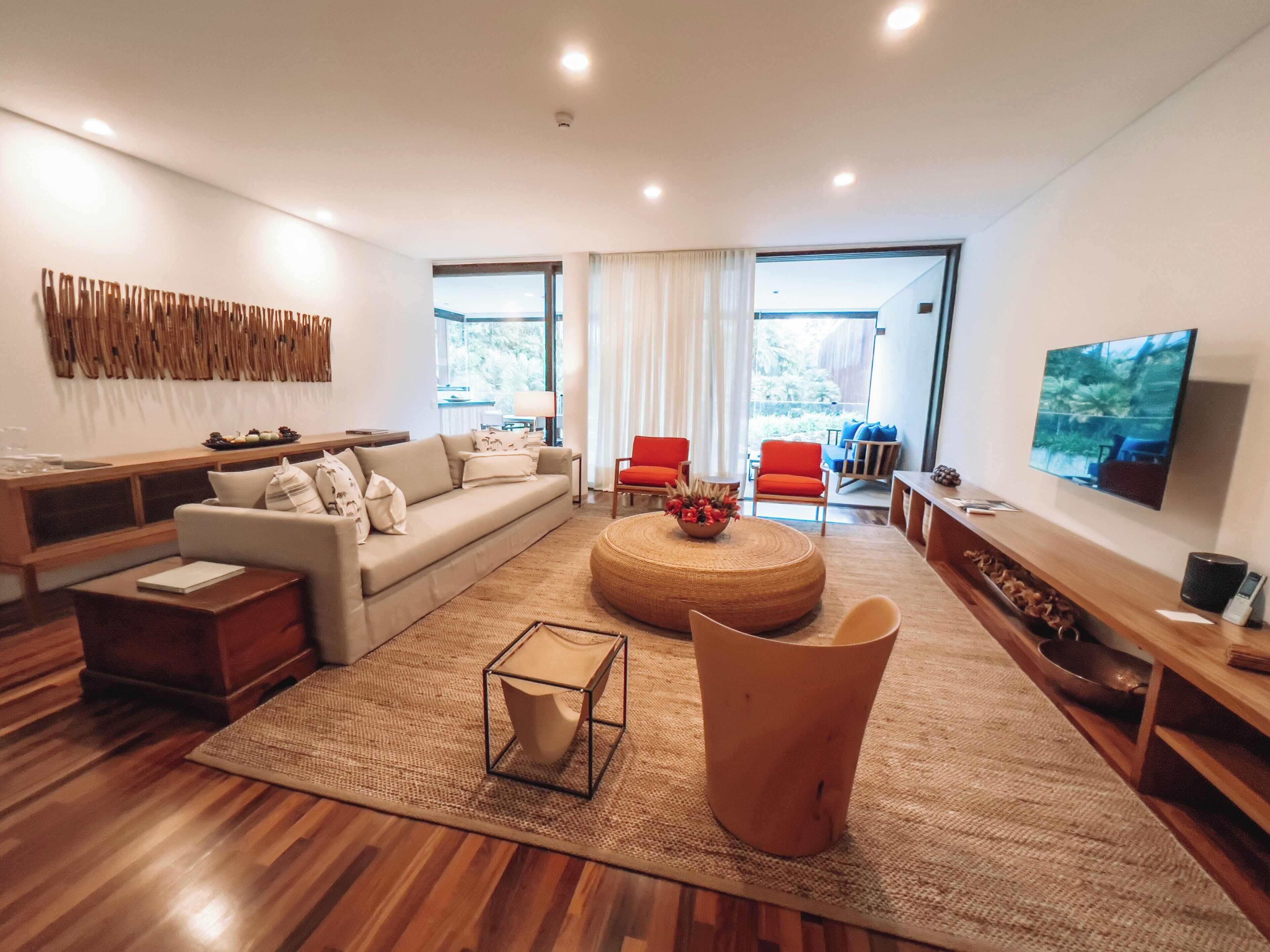 Una sala moderna con sofá, mesa de centro y sillas. Hay un televisor montado en la pared y una ventana que proporciona luz natural.