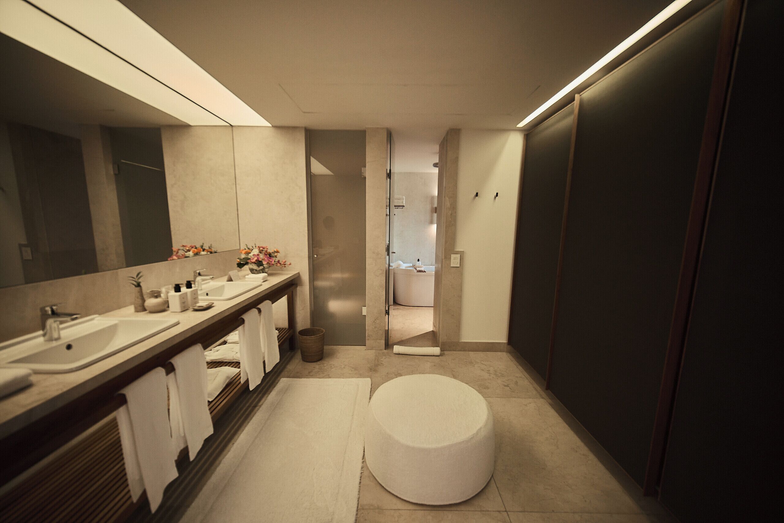 Un baño moderno con un espejo grande, dos lavamanos, un inodoro y un banco redondo.