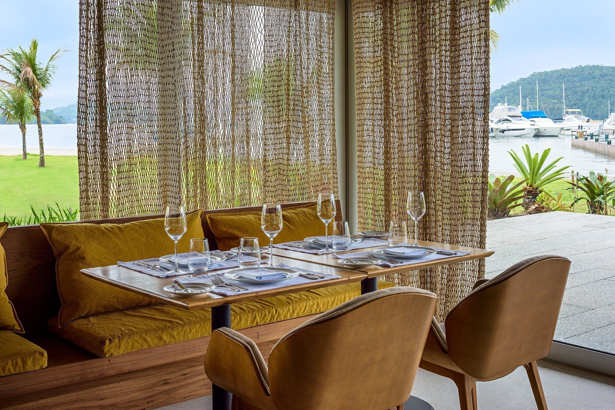 A dining area with a table set for two, a woven partition, and a view of boats and greenery.