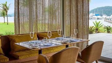 A dining area with a table set for two, a woven partition, and a view of boats and greenery.