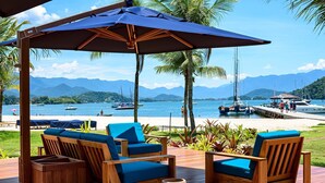A wooden deck with two blue cushioned chairs and a wooden table, overlooking a beach with boats and mountains in the background.