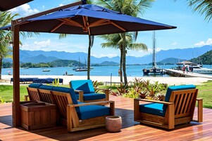 Um deck de madeira com duas cadeiras azuis acolchoadas e uma mesa de madeira, com vista para uma praia e montanhas ao fundo.