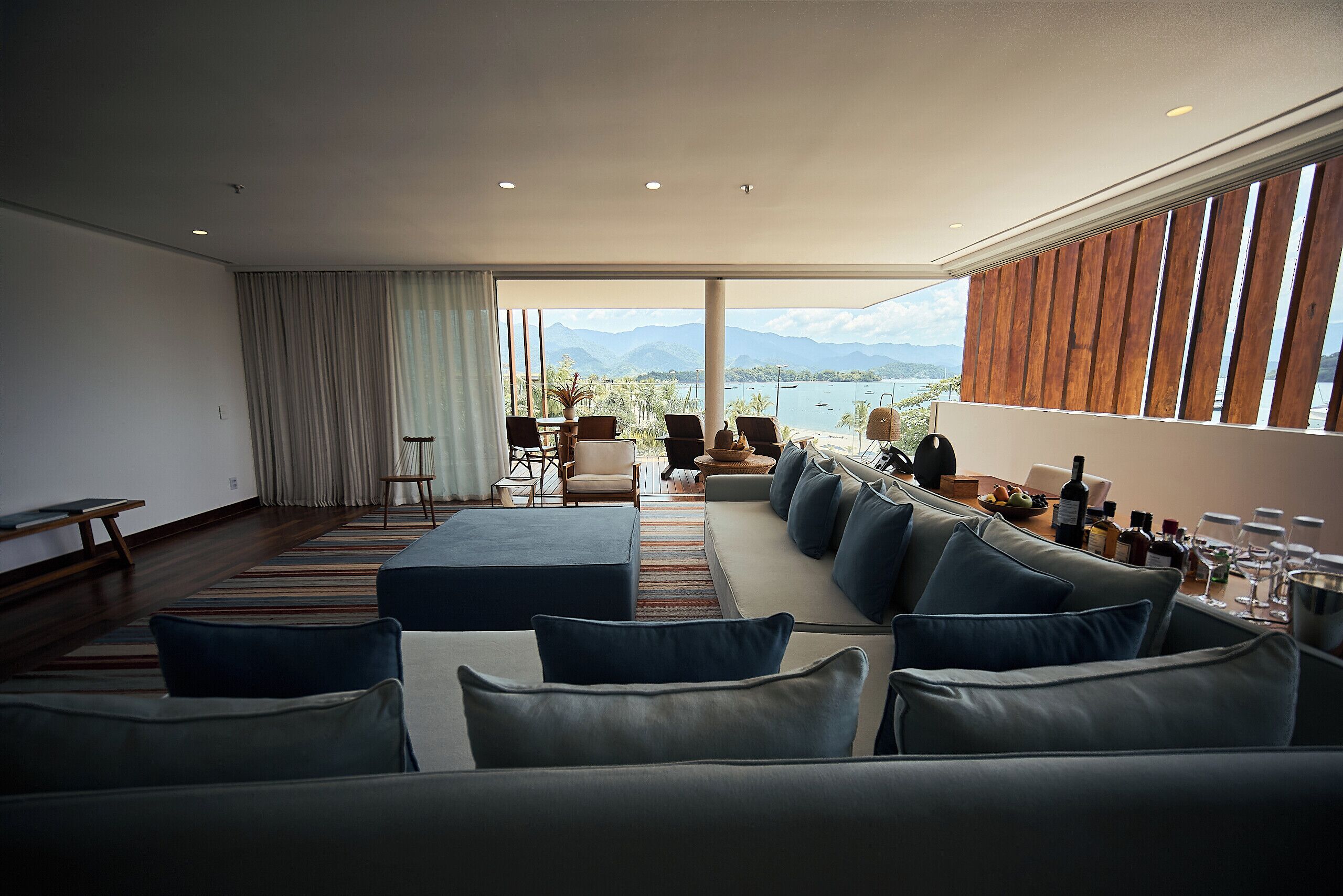 Un salon moderne doté d’un grand canapé, d’un coin repas avec une table dressée pour deux personnes, et offrant une vue sur les montagnes et l’eau grâce à de grandes fenêtres.