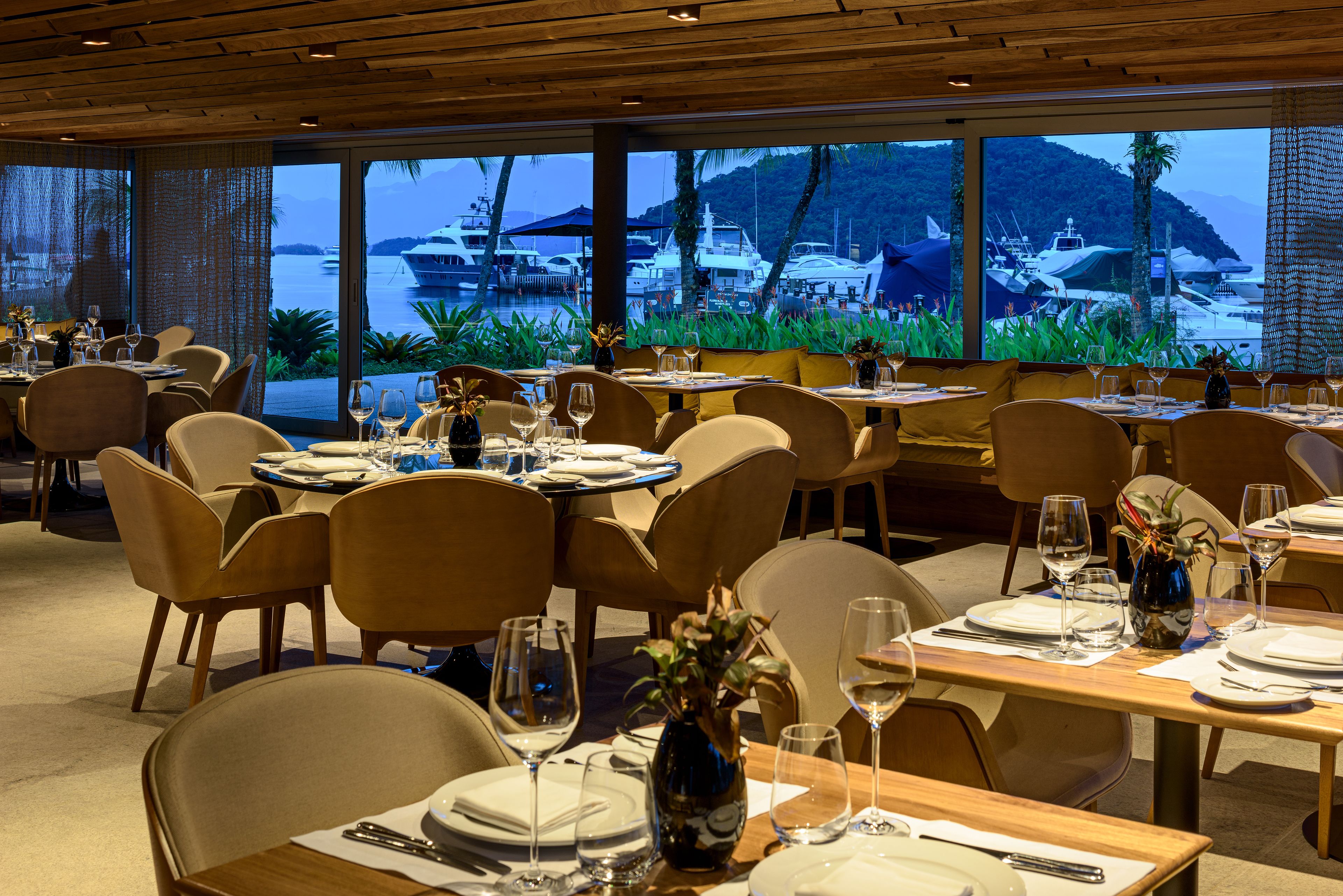 A restaurant with tables set for dining, large windows offering a view of boats and a mountainous landscape.