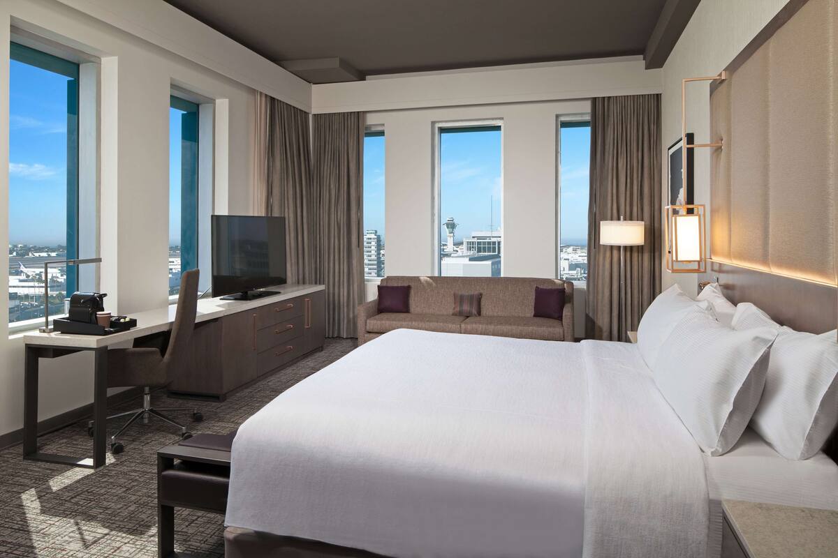 Une chambre d’hôtel dotée d’un grand lit, d’un bureau avec une télévision, d’un canapé et de grandes fenêtres offrant une vue sur la ville.