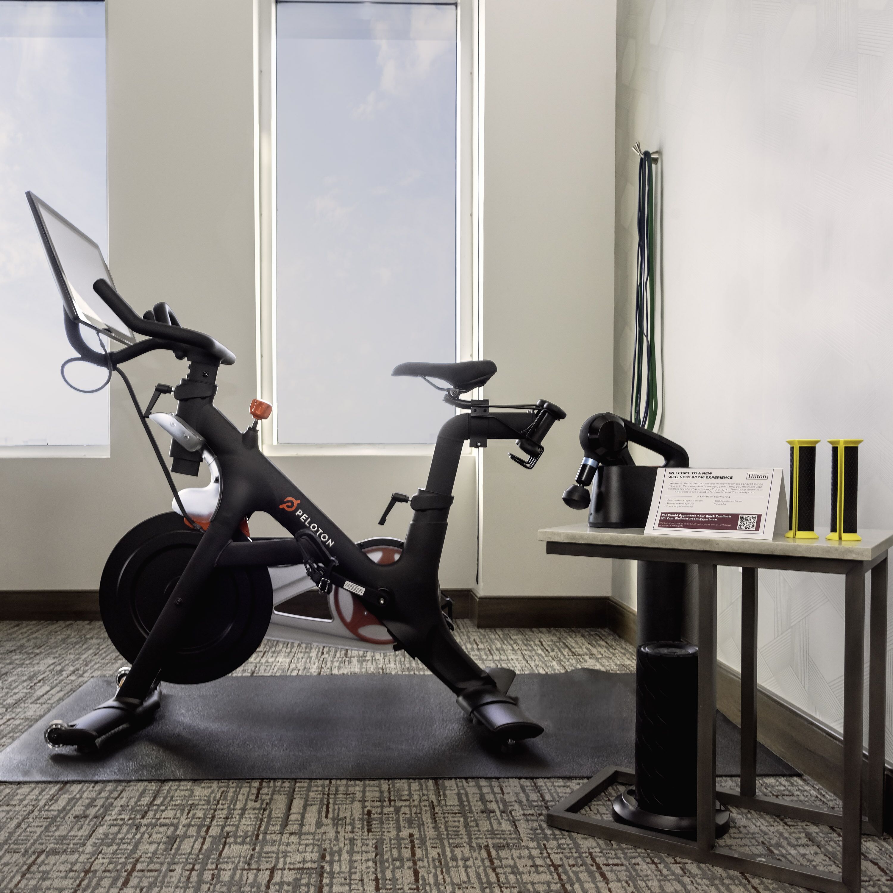 A Peloton stationary bike in a hotel room with a view of the sky through the window.