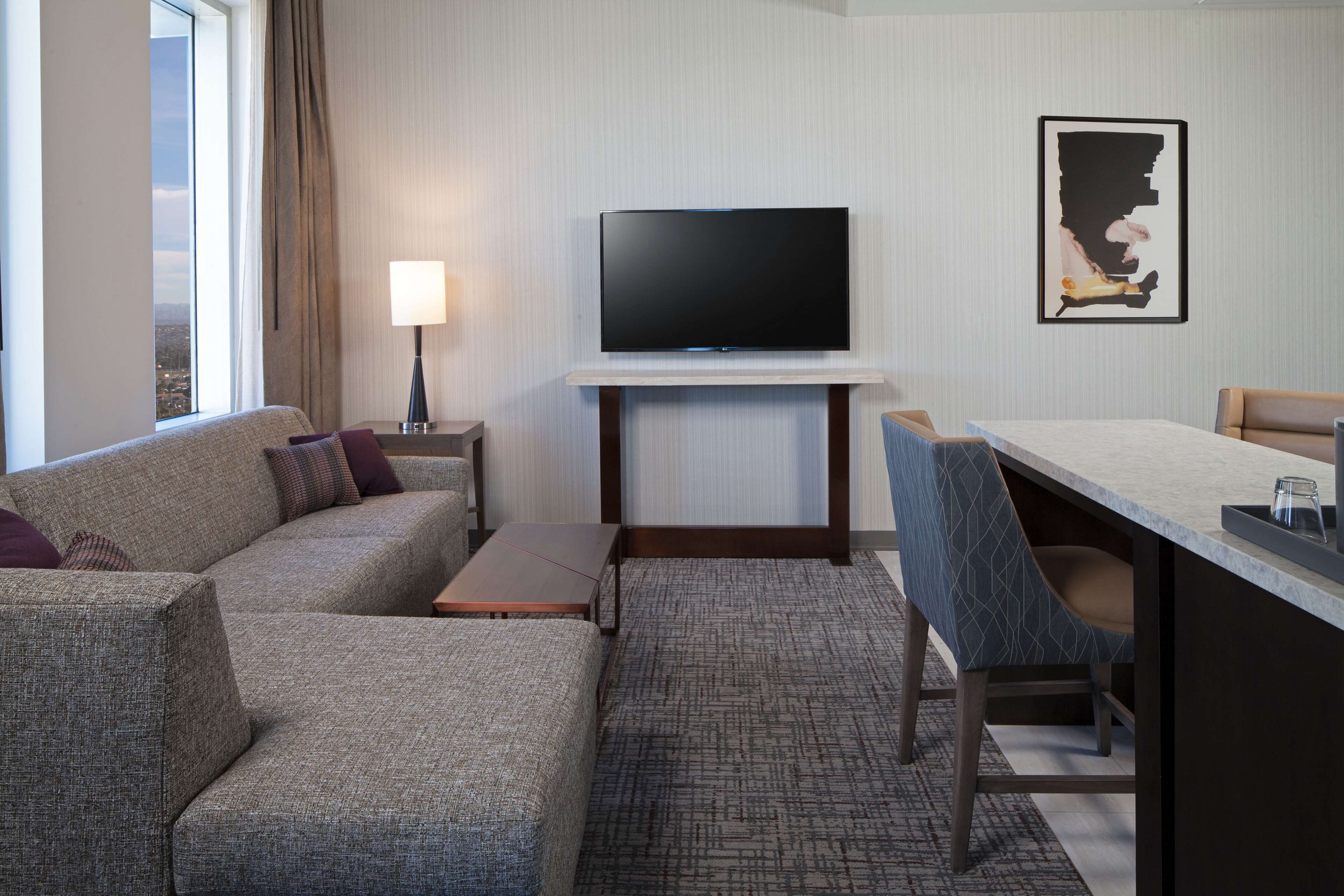 A hotel room with a sofa, a TV mounted on a wall, a desk with a chair, and a window with curtains.