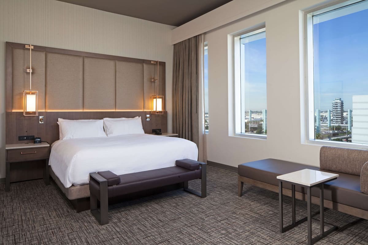 Une chambre d’hôtel avec un grand lit, une table de chevet, une lampe de lecture, un canapé et une fenêtre donnant sur la ville.