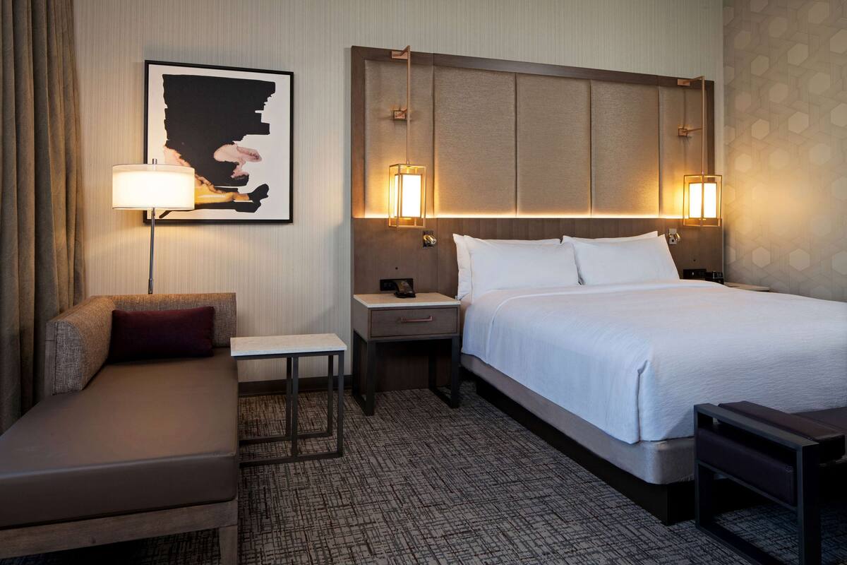 Une chambre d’hôtel avec un lit, un canapé, une petite table et une table de chevet. Un tableau encadré est accroché au mur, et deux lampes sont fixées au mur.