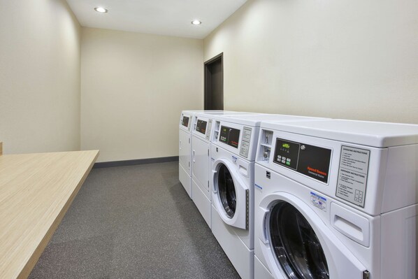 A laundry room with three stacked front-loading washing machines and a wooden table. - Home2 Suites by Hilton Pittsburgh Area Beaver Valley (Monaca)