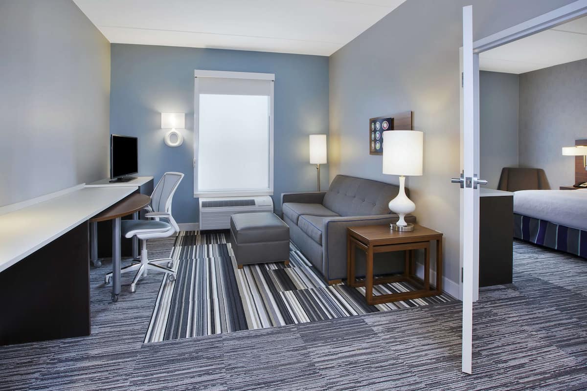Een hotelkamer met een bureau, een sofa, een tafeltje met een lamp, een televisie en een raam met zonwering.