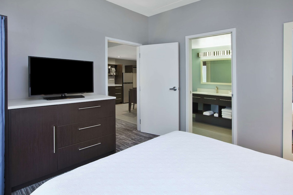 Une chambre d’hôtel avec un lit, un téléviseur posé sur une commode, un dressing et une salle de bain équipée d’un miroir et d’un lavabo.
