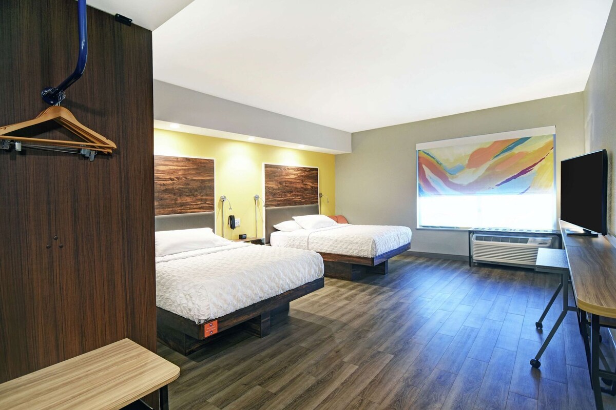A hotel room with two beds, a desk, a TV, and a large abstract painting on the wall.