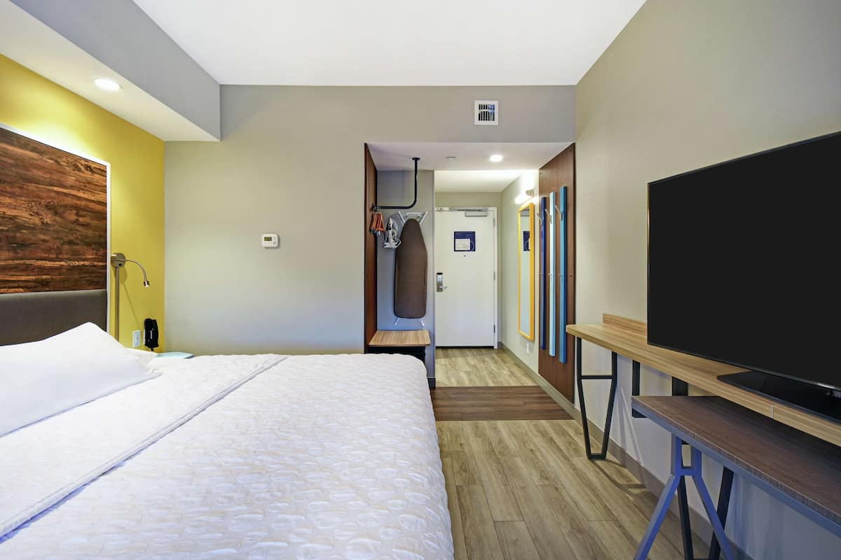 Une chambre d’hôtel dotée d’un grand lit, d’un téléviseur à écran plat, d’un banc en bois, d’un espace douche et d’une porte donnant sur un couloir agrémenté de miroirs.