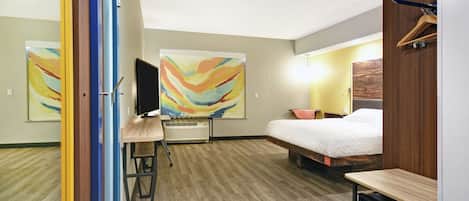 A hotel room with a bed, a desk with a computer, a small fridge, and abstract paintings on the walls.