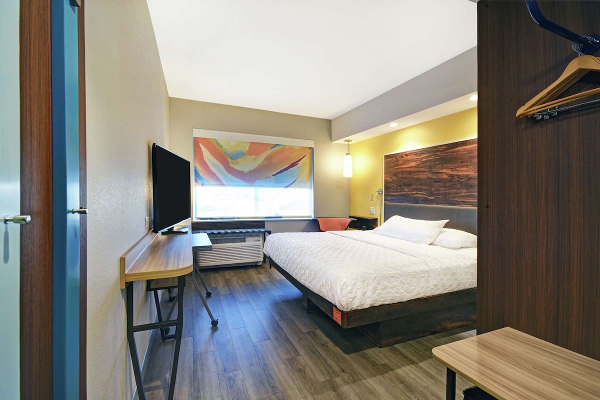 Une chambre d’hôtel comprenant un lit, un bureau avec un téléviseur à écran plat, un radiateur, une fenêtre ornée d’un tableau abstrait aux couleurs vives et une tête de lit en bois.